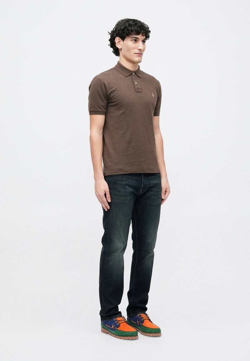 Junger Mann steht in einem braunen kurzärmeligen Polo-Shirt, dunk blauem Jeans und bunten, mehrfarbigen Bootsschuhen vor einem weißen Hintergrund.