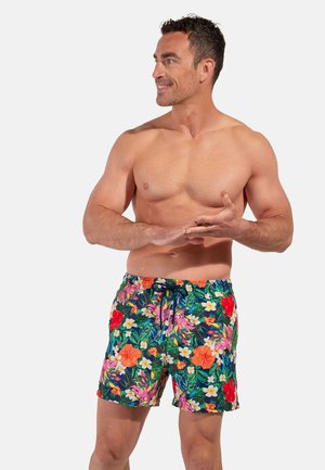 Hombre musculoso de pie sin camiseta, con pantalones cortos de baño florales y coloridos, mirando hacia un lado con las manos entrelazadas al frente.
