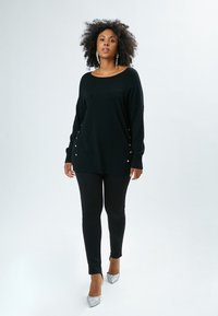 Maglione nero a maglia con bottoni laterali, abbinato a leggings neri e tacchi argentati lucidi. La modella ha i capelli ricci e si trova in piedi davanti a uno sfondo chiaro.