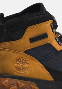 Botas Timberland impermeables con parte superior de ante marrón, acentos de textil negro, un diseño texturizado en azul marino y suela de goma con patrón.