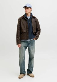 Bărbat tânăr purtând o șapcă albastru deschis, o jachetă maro peste un pulover bleumarin cu fermoar, blugi decolorați și mocasini bej, stând cu o mână în buzunar.