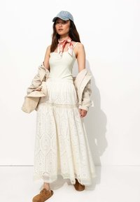 Jupe maxi blanche à motifs avec des détails en œillets, assortie à un débardeur léger, une veste beige, une écharpe rouge et une casquette en denim. Chaussures brunes à enfiler.