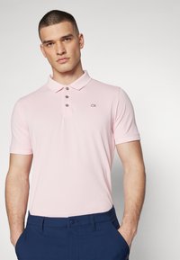 Šviesiai rožinė polo marškinėliai, trumpomis rankovėmis, su dviem mygtukais, CK logotipas ant krūtinės, derinami su tamsiai mėlynomis kelnėmis, glotni tekstūra.