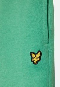 Green fabric with a side pocket and a small embroidered yellow bird emblem outlined in black near the pocket seam.