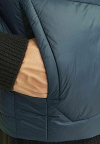 Jachetă puffer bleumarin, material matlasat, cu o mână în buzunarul lateral. Jacheta are o textură netedă și o ușoară strălucire.