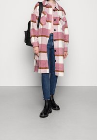 Lång rosa och vit rutig kappa med bruna ränder, knäppt fram, två bröstfickor, i kombination med blåjeans och svarta ankelskor.