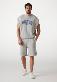 Siva dukserica kratkih rukava s plavim "GUESS" logotipom, u kombinaciji s odgovarajućim šortsevima. Ima ležeran kroj i mekanu teksturu. Pobjednički izgled upotpunjuju bijele tenisice.
