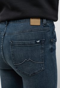 Jeans de mezclilla azul oscuro con un ligero desgaste, una etiqueta de cuero de Mustang en la cintura y un distintivo bordado en los bolsillos traseros.