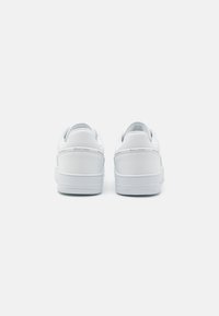 Bijele tenisice s glatkom kožnom površinom, podstavljenim petama i gumenim potplatom. Minimalistički dizajn bez vidljivih logotipa ili uzoraka.