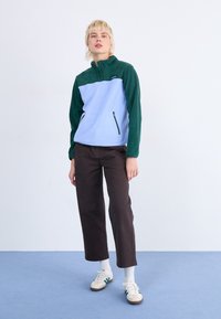 Fleece pullover in light blue and green, features a high collar, side zip pocket, and ribbed cuffs. Paired with dark trousers and sneakers.