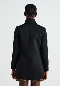 Černý přizpůsobený kabát s vysokým límcem, dlouhými rukávy a strukturovaným designem. Nosený přes sukni s leopardím potiskem, která vykazuje texturovanou tkaninu.