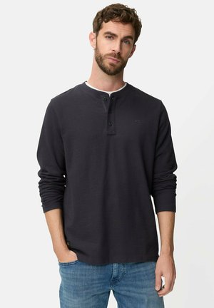 Man met baard, gekleed in een zwarte henley met lange mouwen en een blauwe spijkerbroek, staande met één hand in zijn zak tegen een effen grijze achtergrond.