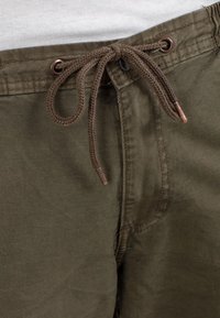 Close-up of olive green pants with a tied brown drawstring and visible stitching against a white fabric background.