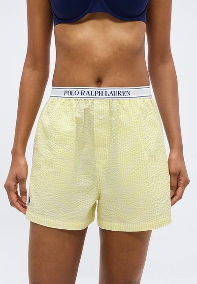 Γυναίκα με κίτρινο καρό μποξεράκι Polo Ralph Lauren με λευκό ζωνάρι στη μέση και μπλε σουτιέν, εμφανίζεται από το ύψος της μέσης έως το μέσο των μηρών.