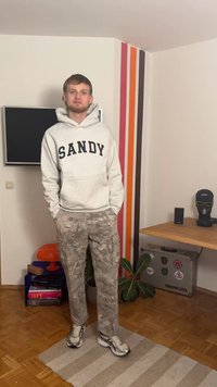 Sivi duks s kapuljačom s natpisom "SANDY" u crnim slovima, maskirne cargo hlače i srebrne tenisice. U pozadini se vidi drveni pod i šarene prugaste zidove.