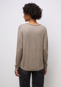 Donna con capelli ricci corti, indossa un maglione beige a maniche lunghe e jeans neri, rivolta verso l'asse contro uno sfondo chiaro e semplice.