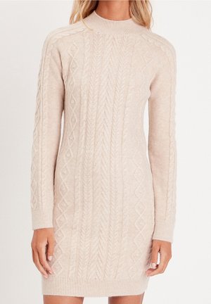 Femme portant une robe-pull beige à manches longues en maille avec des motifs torsadés et en losanges, des poignets et un ourlet côtelés.