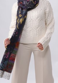 Personne portant un pull crème en maille torsadée, un pantalon beige et une écharpe foncée à motif floral avec des franges, tenant les extrémités de l'écharpe.