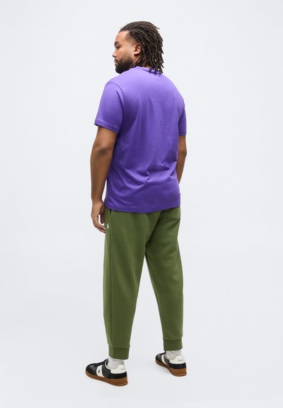 Άνδρας με dreadlocks που φοράει μοβ κοντομάνικη μπλούζα, παντελόνι λαδί και μαύρα-λευκά αθλητικά παπούτσια, στέκεται γυρισμένος πλάτη σε λευκό φόντο.