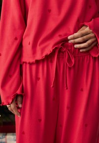 Punane puhkekostüüm, mis sisaldab pikka varrukat ja pükse. Kanga materjal on pehme ning sellele on trükitud väikesed südame mustrid. Äär on laineline ning vöökoht on reguleeritav paelaga.