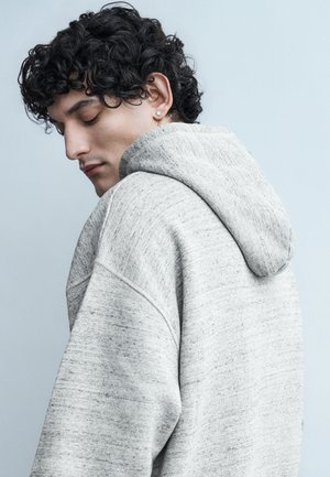 Νεαρός άνδρας με σγουρά μαλλιά, που φοράει γκρι ανοιχτό φούτερ με κουκούλα, κοιτάζει κάτω και είναι ελαφρώς γυρισμένος προς τα πλάγια, με φόντο απλή επιφάνεια.