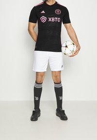 Musta jalkapallopaita, jossa vaaleanpunaiset yksityiskohdat, valkoiset shortsit, mustat sukat valkoisilla raidoilla ja mustat nappikengät, pitelee moniväristä jalkapalloa.