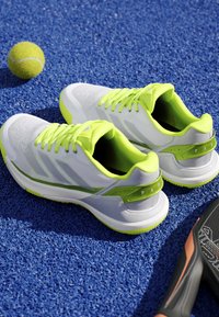 White and neon green tennis shoes on blue court surface with tennis ball and part of black tennis racket visible.