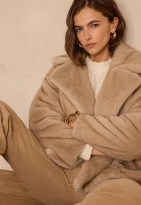 Manteau en fausse fourrure beige avec un large col, porté sur un pull en maille crème. Pantalon en velours côtelé marron et bracelets dorés visibles. Fond minimaliste.