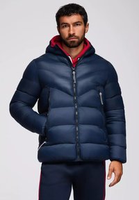 Giubbotto imbottito blu navy con cappuccio, caratterizzato da cuciture a zigzag, due tasche laterali con zip e un colletto foderato in rosso per maggiore calore.