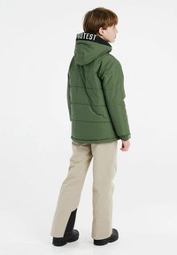 Giacca a vento verde con cappuccio e colletto nero, caratterizzata da cuciture orizzontali. Indossata con pantaloni beige e stivali neri.