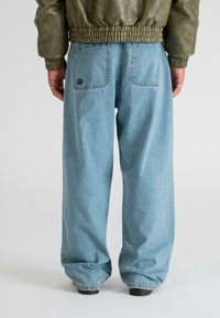 Jeans de mezclilla de corte recto en azul claro con textura desgastada y bolsillos traseros. Combinados con una chaqueta de cuero verde oliva.