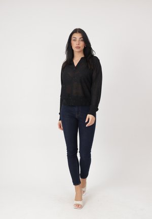 Kobieta o długich ciemnych włosach, ubrana w przezroczystą czarną bluzkę z długim rękawem, ciemne obcisłe dżinsy i białe buty na obcasie z odkrytymi palcami, stojąca na tle jednolitej białej ściany.