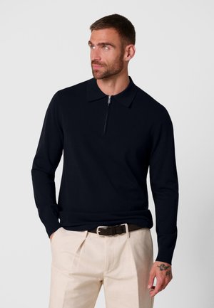 Hombre con una camisa polo negra de manga larga con cremallera y pantalones beige con cinturón negro, mirando hacia su izquierda con las manos en los bolsillos.