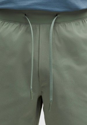 Detailní záběr zelených kalhot se stahovací šňůrkou, měkkou látkovou texturou a viditelným pasem pod modrým topem.