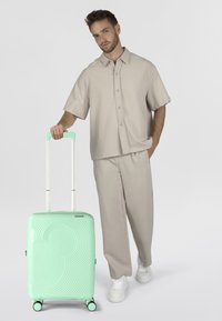 Valise à coque rigide vert menthe avec un motif ondulé, poignée rétractable argentée et quatre roues noires. Présentée à côté d'une personne vêtue de tenue neutre.