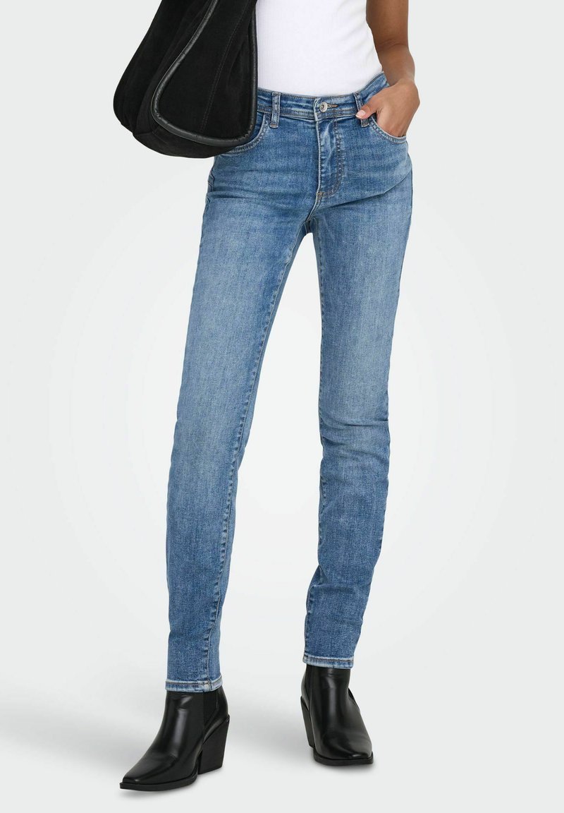 Person iført lyseblå skinny jeans, svarte ankelstøvler, hvit topp, bærer stor svart skulderveske, står med en hånd i lommen.