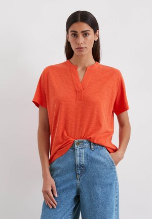 T-shirt à manches courtes et col en V de couleur orange vif, en tissu léger. Présente une coupe décontractée et un détail noué à l'avant. Porté avec un jean en denim bleu.