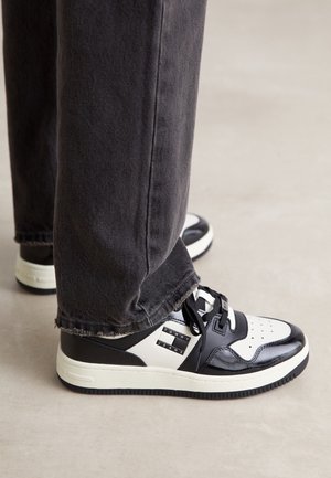 Personne portant des baskets Tommy Jeans noires et blanches et un pantalon gris foncé, debout sur un sol clair.