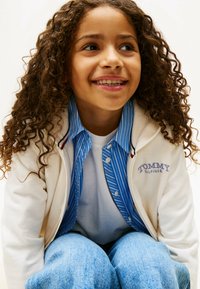 Biała kurtka na zamek błyskawiczny, niebieska koszula w paski pod spodem, biała koszulka, dżinsy. Cechy: kręcone włosy i uśmiech na twarzy.