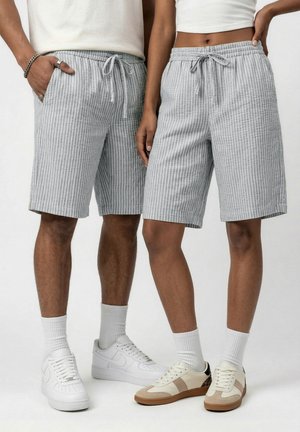 Dos personas con pantalones cortos de rayas verticales en gris claro con cordón, medias blancas y zapatillas, de pie uno al lado del otro.