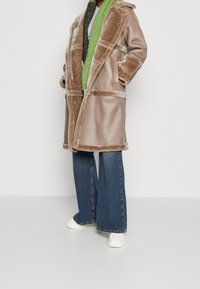 Beige faux fur coat with a wide collar, large pockets, and a tie waist, paired with a green textured scarf and blue wide-leg jeans.