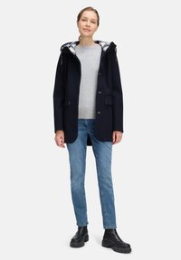 Kvinde står iført en navy hættejakke over en grå sweater, lyseblå jeans og sorte klodsede støvler mod en ensartet lys baggrund.