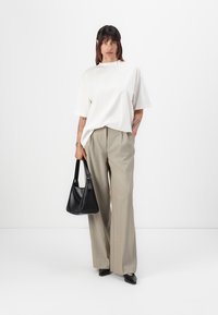 Hvit oversized t-skjorte, beige bred-bein bukser, svart veske. Modell står med synlige tatoveringer, mot en enkel hvit bakgrunn.