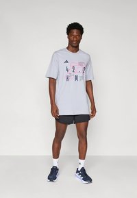 Sivá športová tričko s čiernym textom a ružovými akcentmi, s krátkymi rukávmi a voľným strihom, v kombinácii s čiernymi šortkami a športovou obuvou.