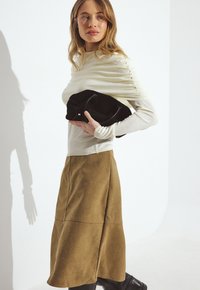 Kreemjas tipis ja pruunis seelikus naine hoiab musta kotti valge tausta ees, vaadates veidi vasakule.