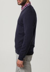 Marineblaues Sweatshirt mit Rundhalsausschnitt, aus weichem Stoff, kombiniert mit einem rot-blauen karierten Hemd darunter. Graue Hose.