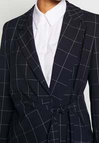 Marinblå blazer med vitt rutmönster, med ett bälte i midjan, snedskurna slag och en strukturerad design. Under finns en vit skjorta med krage.