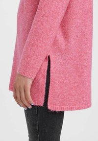 Pörröinen vaaleanpunainen neulepaita pitkillä hihoilla, sivuhalkioilla ja ribsikujilla. Yhdistetty tummansiniset farkut, jotka esittelevät rentoa tyyliä.