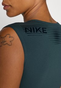 Top sportiv fără mâneci de culoare verde închis, cu un design texturat pe spate ce include logo-ul "NIKE" și linii orizontale, realizat dintr-un material neted și elastic.