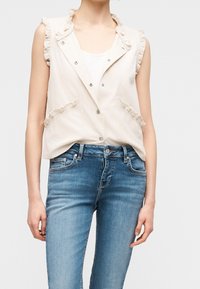 Gilet beige sans manches avec bords froncés porté sur un haut blanc, associé à un jean bleu ajusté sur une personne debout.